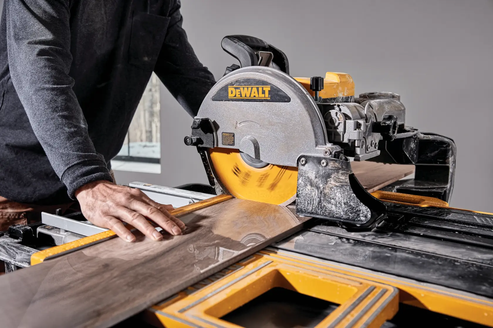 A person operating a DEWALT D36000-QS wet tile saw in a well-lit room, cutting a large tile with water splashing on the work surface.