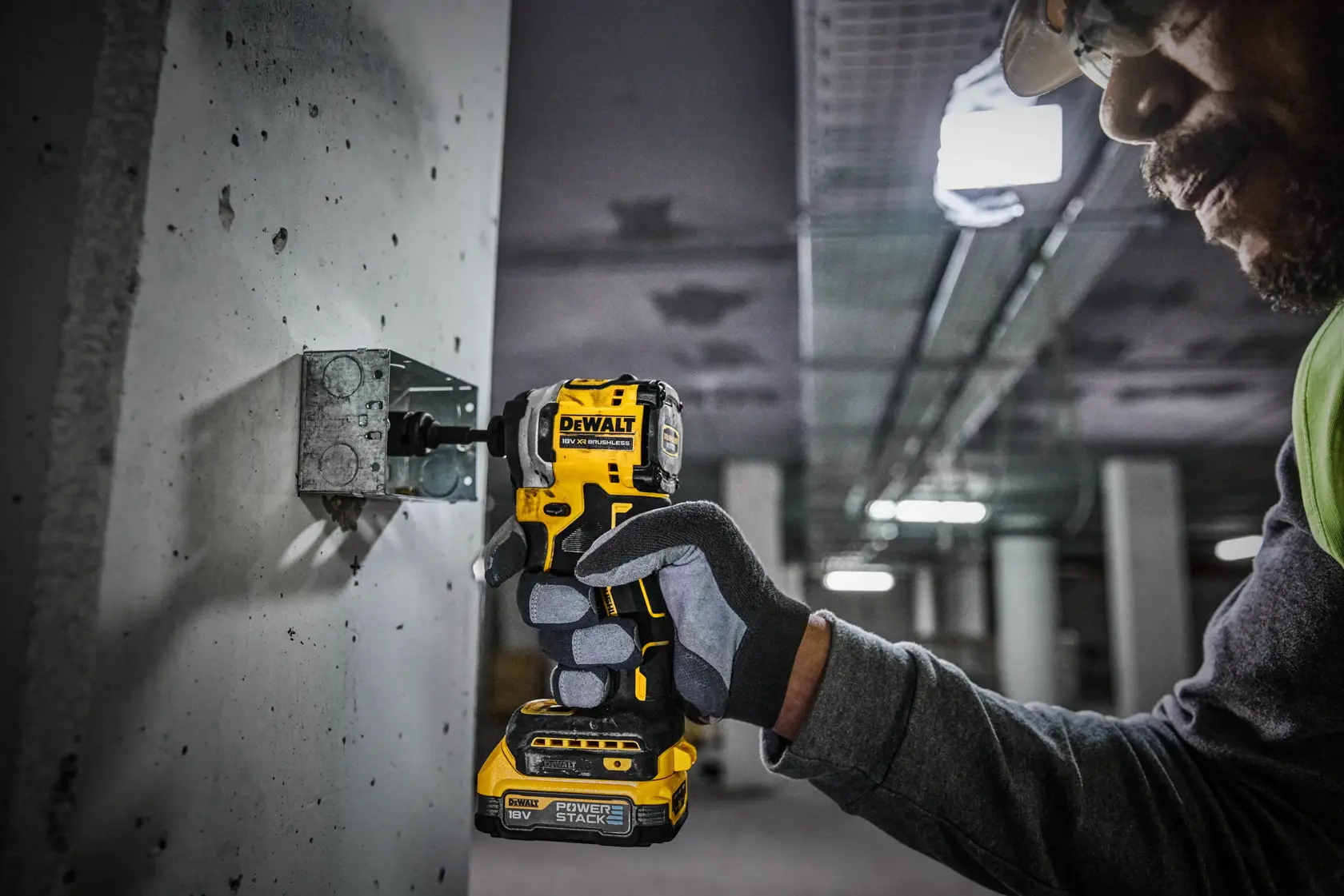 A person wearing gloves uses a DEWALT 18V cordless impact driver to fasten a metal bracket onto a concrete surface in an industrial setting.