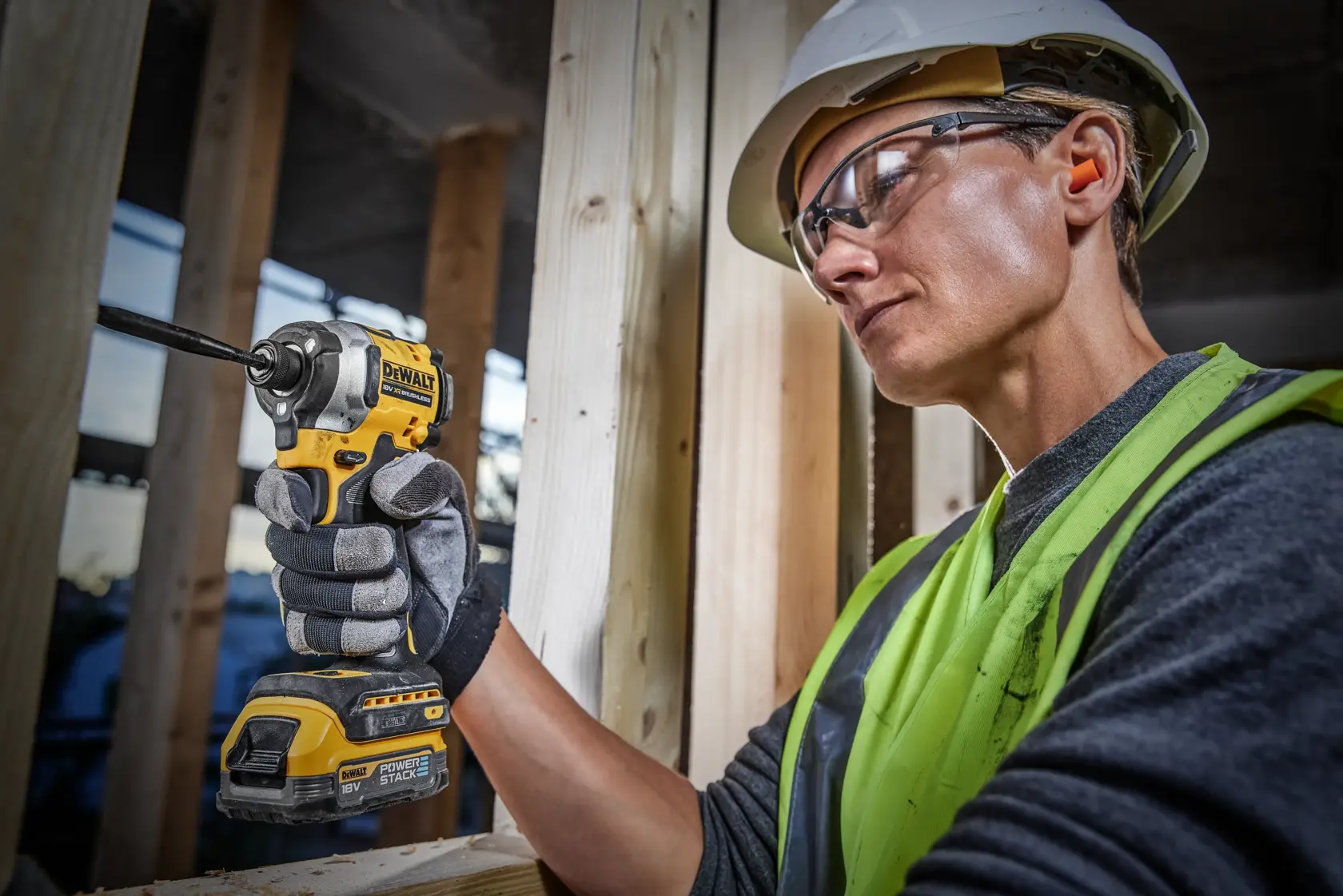 A person wearing a safety helmet and high-visibility vest is using a DEWALT DCF850E1T cordless impact driver to fasten a screw into wooden beams at a construction site. Their face is blurred for privacy.