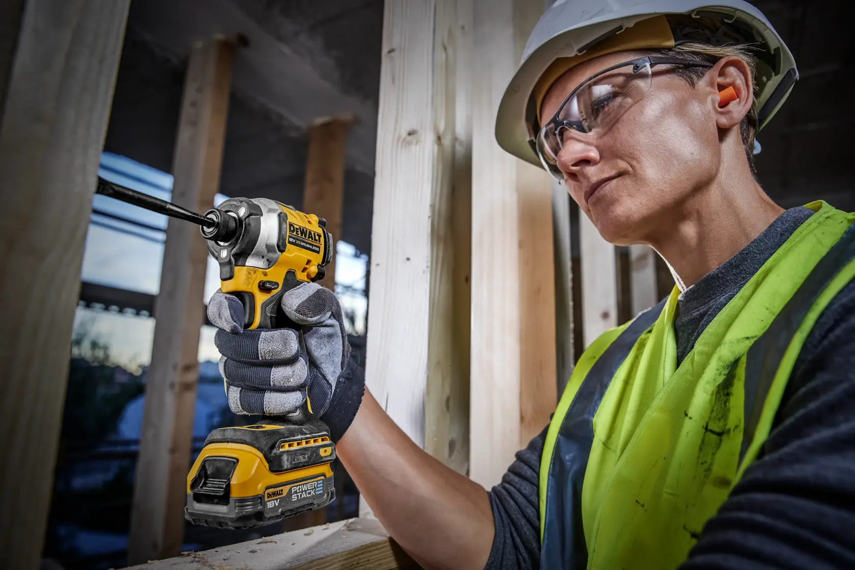 A person wearing a safety vest, gloves, and a hard hat is using a DEWALT DCF850E1T cordless impact driver at a construction site, holding the tool against a wooden frame.