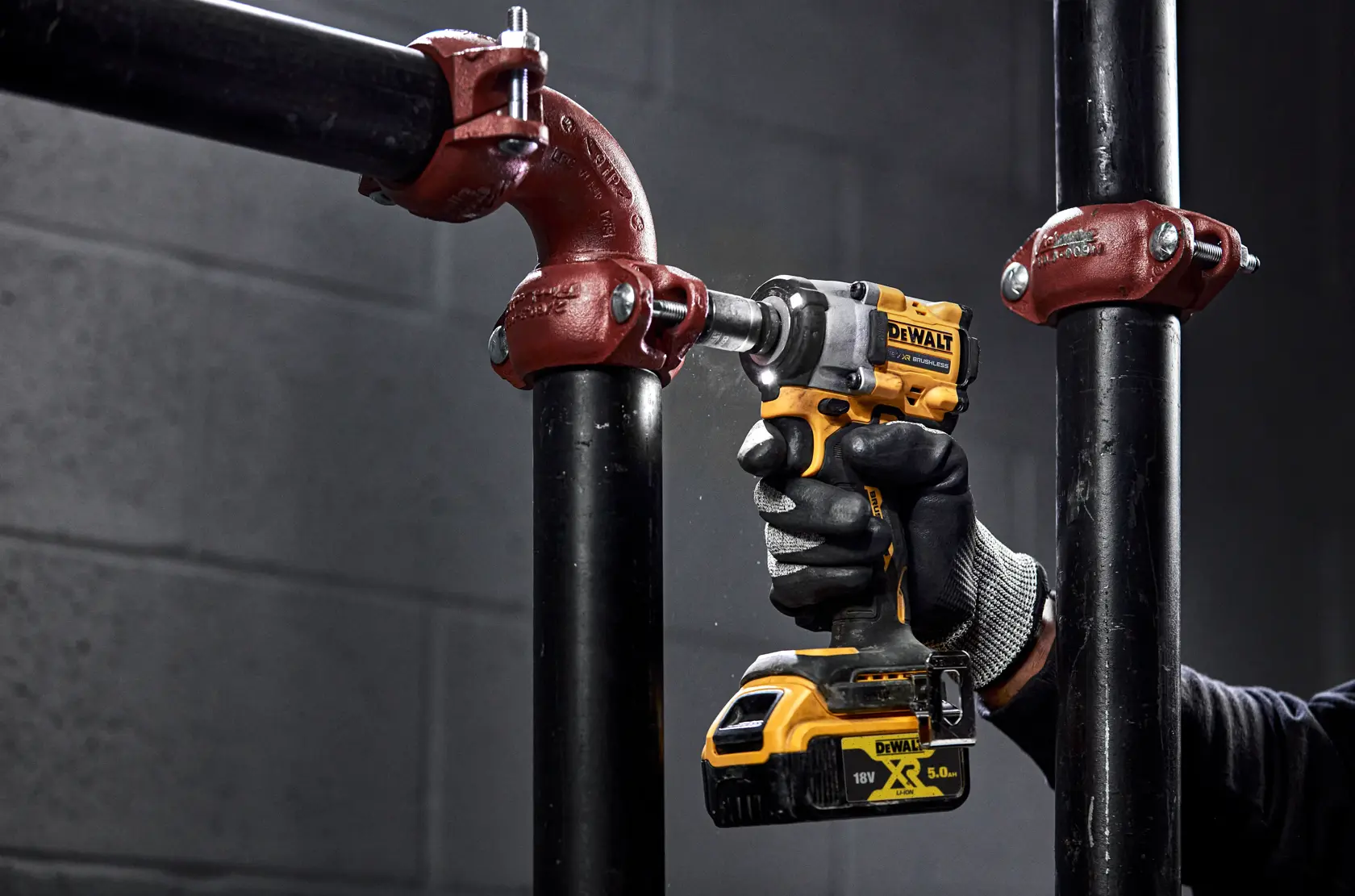 A person using a yellow and black DEWALT cordless impact wrench to work on a metal pipe joint, wearing black gloves. The tool is being used to tighten or loosen bolts in an industrial setting with black pipes and red pipe fittings.