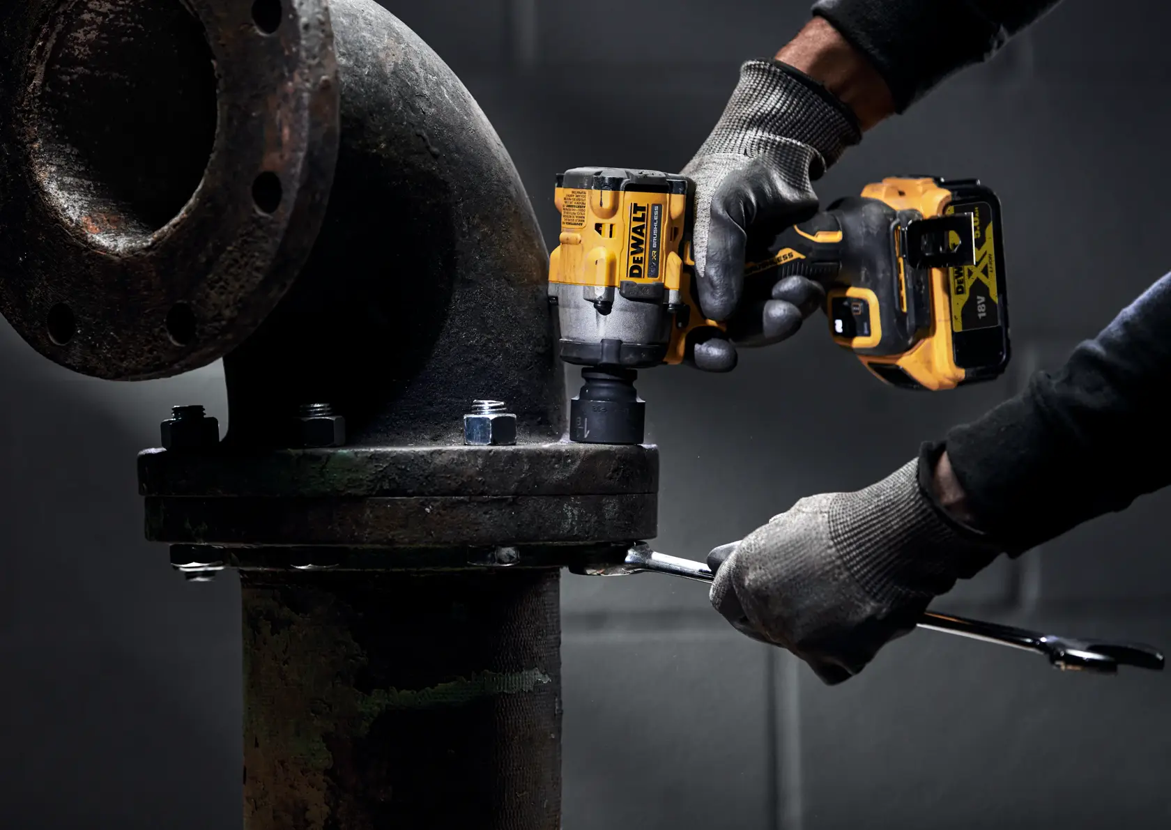 A person wearing gloves uses a DEWALT cordless impact wrench (DCF923N) on a large metal pipe flange, tightening bolts in an industrial setting.