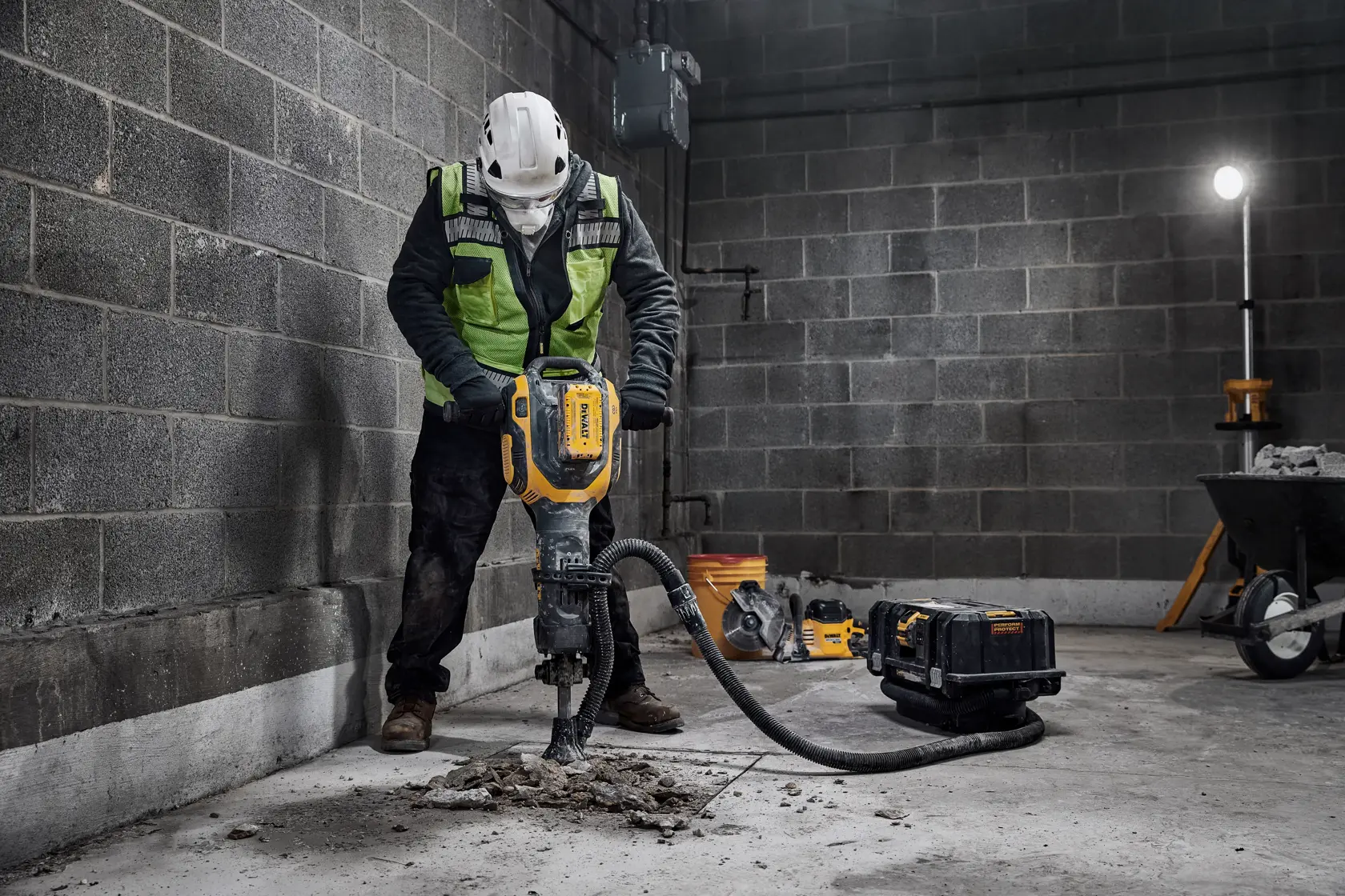 Un trabajador de la construcción con equipo de seguridad maneja un martillo demoledor eléctrico DEWALT para romper hormigón en una obra. La herramienta está conectada a un sistema de aspiración y hay equipos de construcción al fondo.