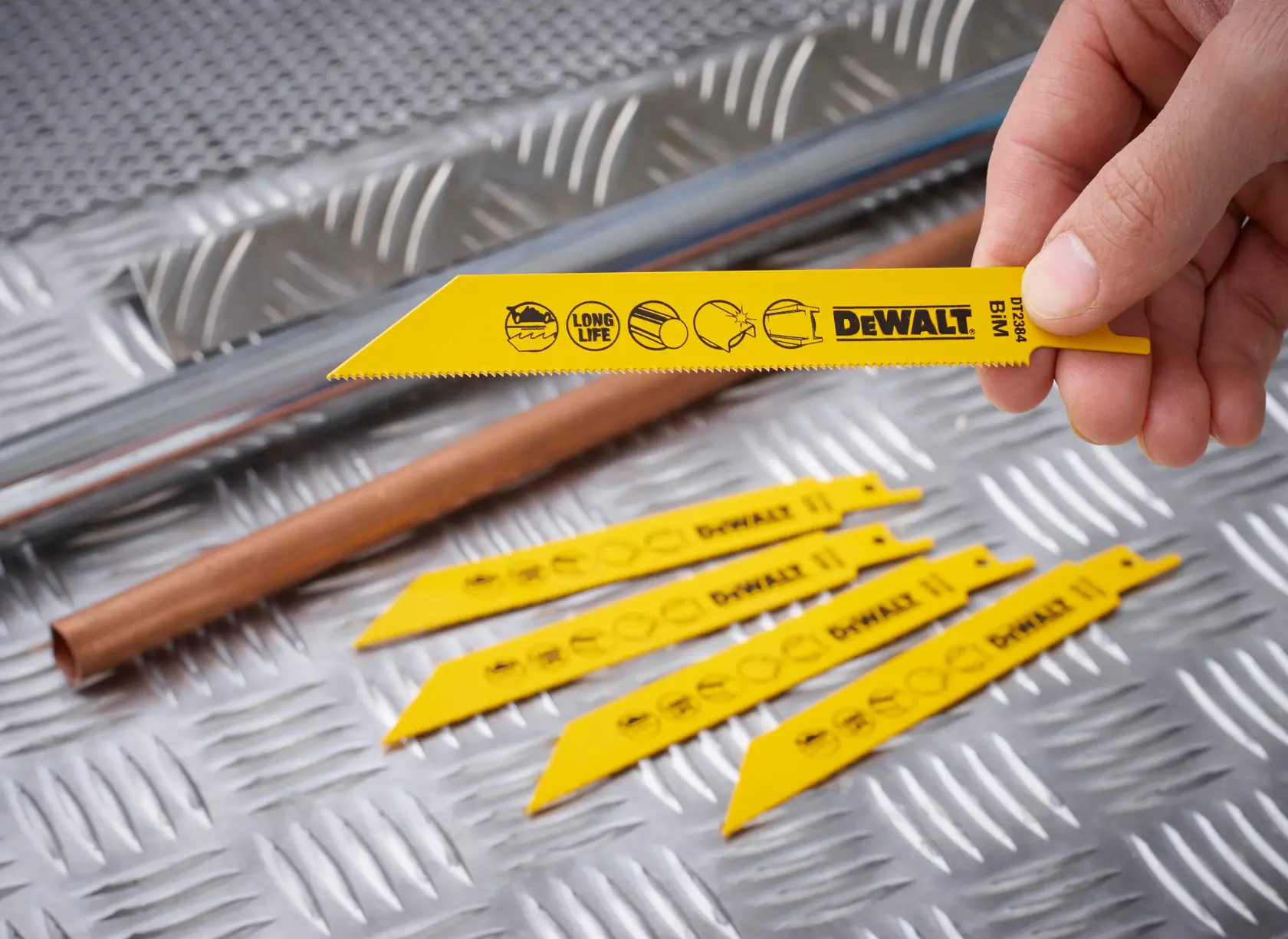 Una mano sostiene una hoja DEWALT DT2384 amarilla y otras están sobre chapa metálica junto a tubos.