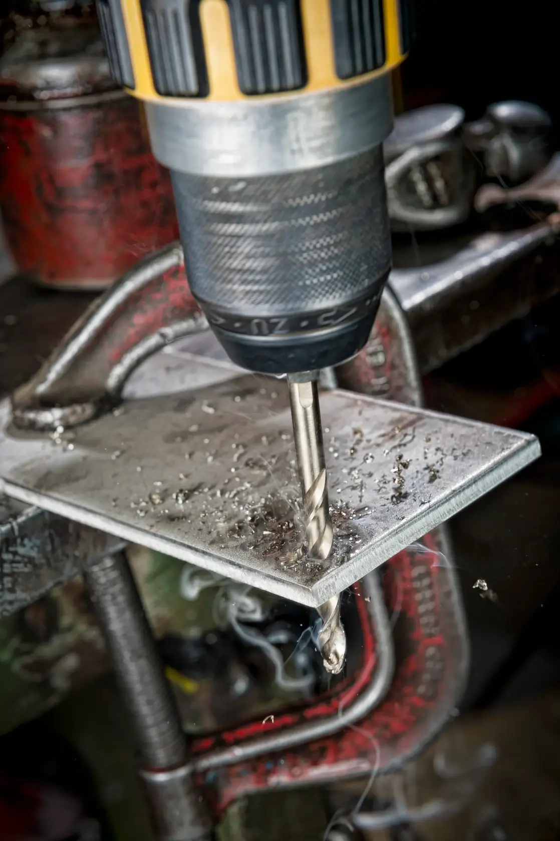 A drill bit makes a hole in a metal sheet held by a clamp, with metal shavings scattered around the area.