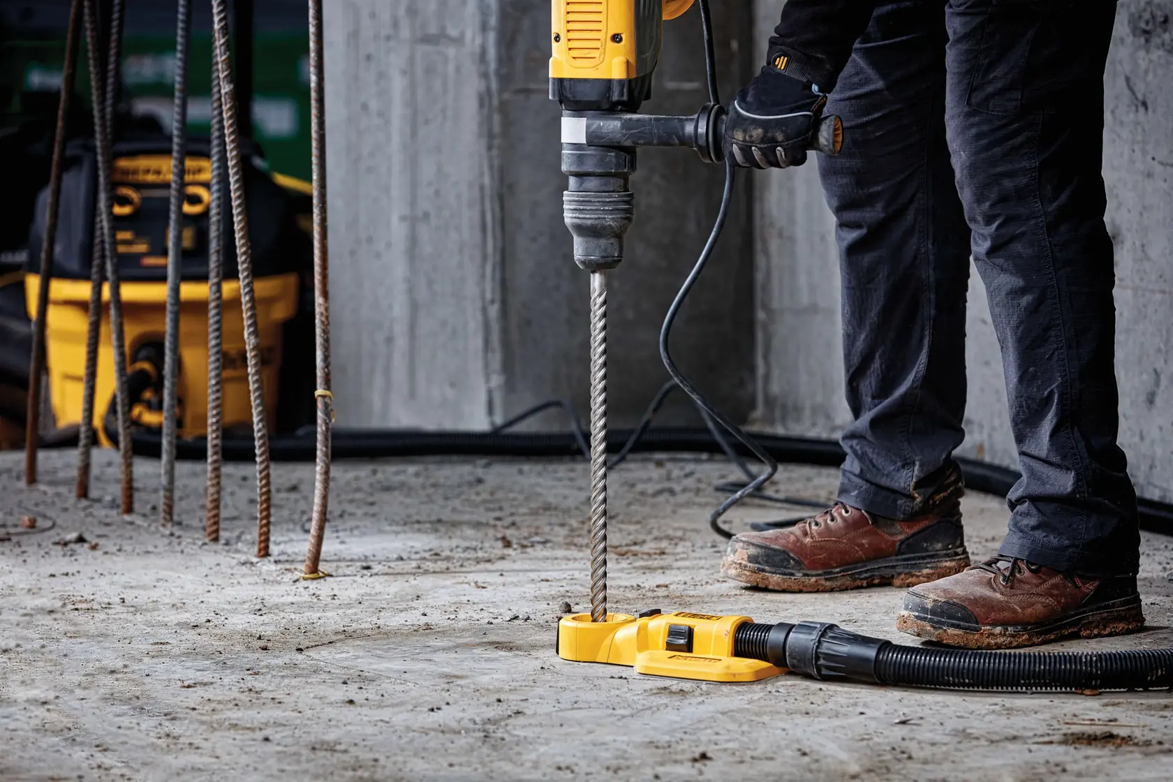 Eine Person mit Arbeitshandschuhen und -stiefeln verwendet einen gelben DEWALT-Bohrhammer, um einen Betonboden zu bohren. Die Bohrmaschine ist an ein Staubabsaugsystem angeschlossen. Metallische Bewehrungsstäbe und eine DEWALT Werkzeugtasche sind im Hintergrund zu sehen.