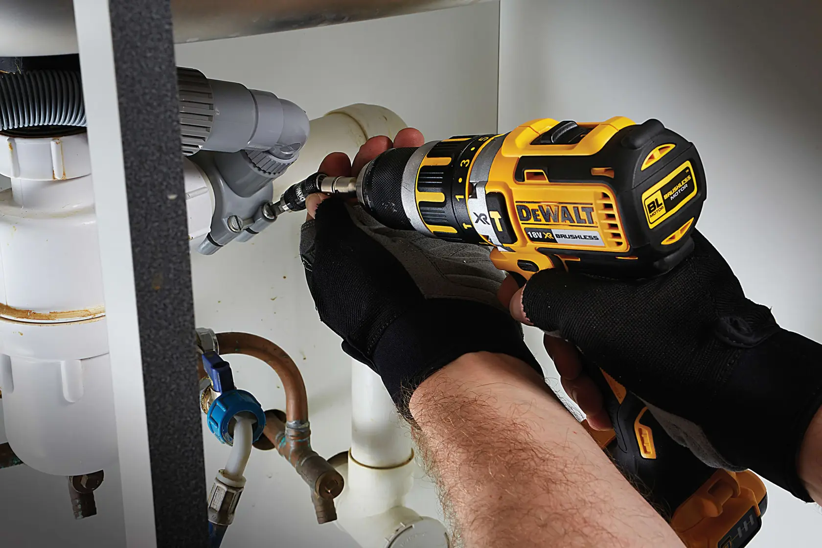 Persona usa taladro DEWALT en tuberías bajo un lavabo, con guantes negros de seguridad.