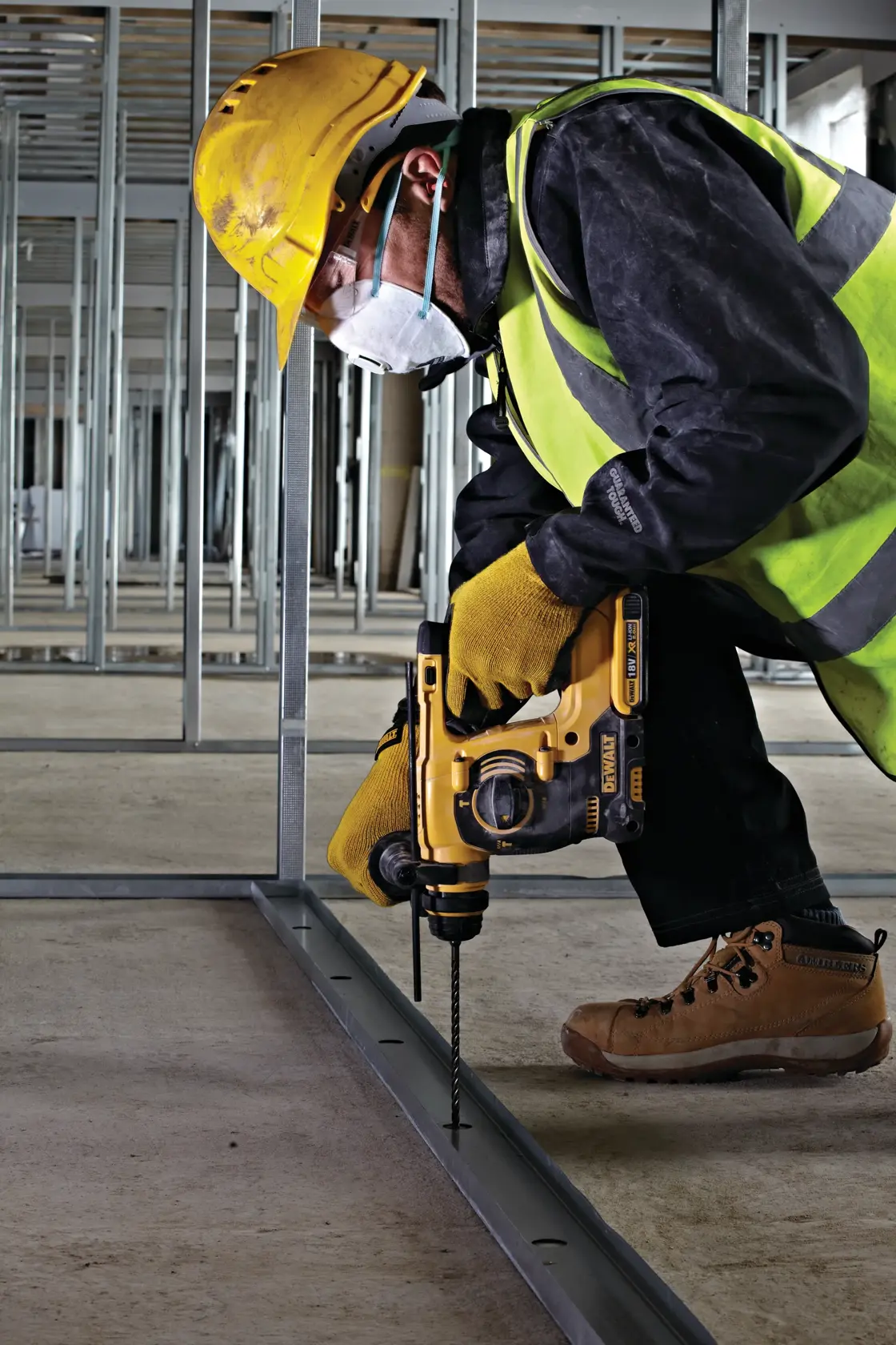 Trabajador usa taladro DEWALT en obra, lleva equipo de seguridad y perfora marco metálico en el suelo.