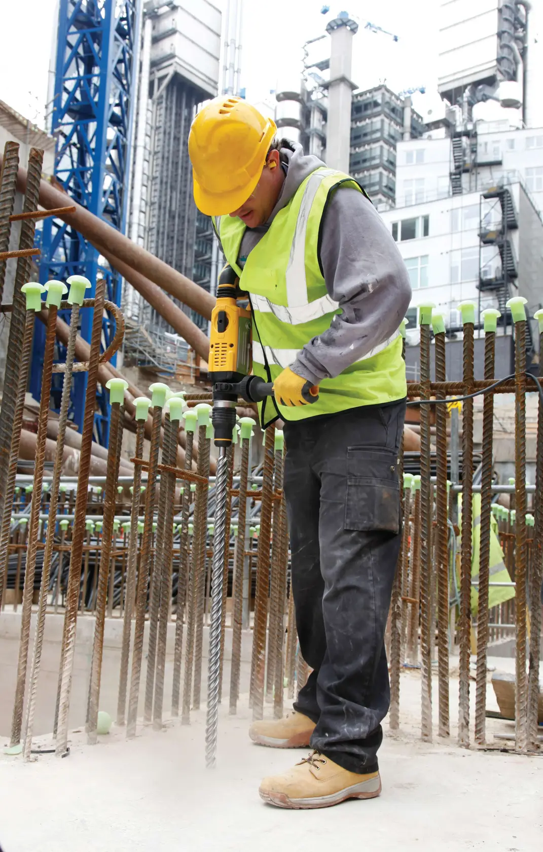 Arbeiter nutzt DEWALT-Bohrmaschine auf Baustelle, mit Sicherheitskleidung zwischen Stahlstangen und Gebäuden.