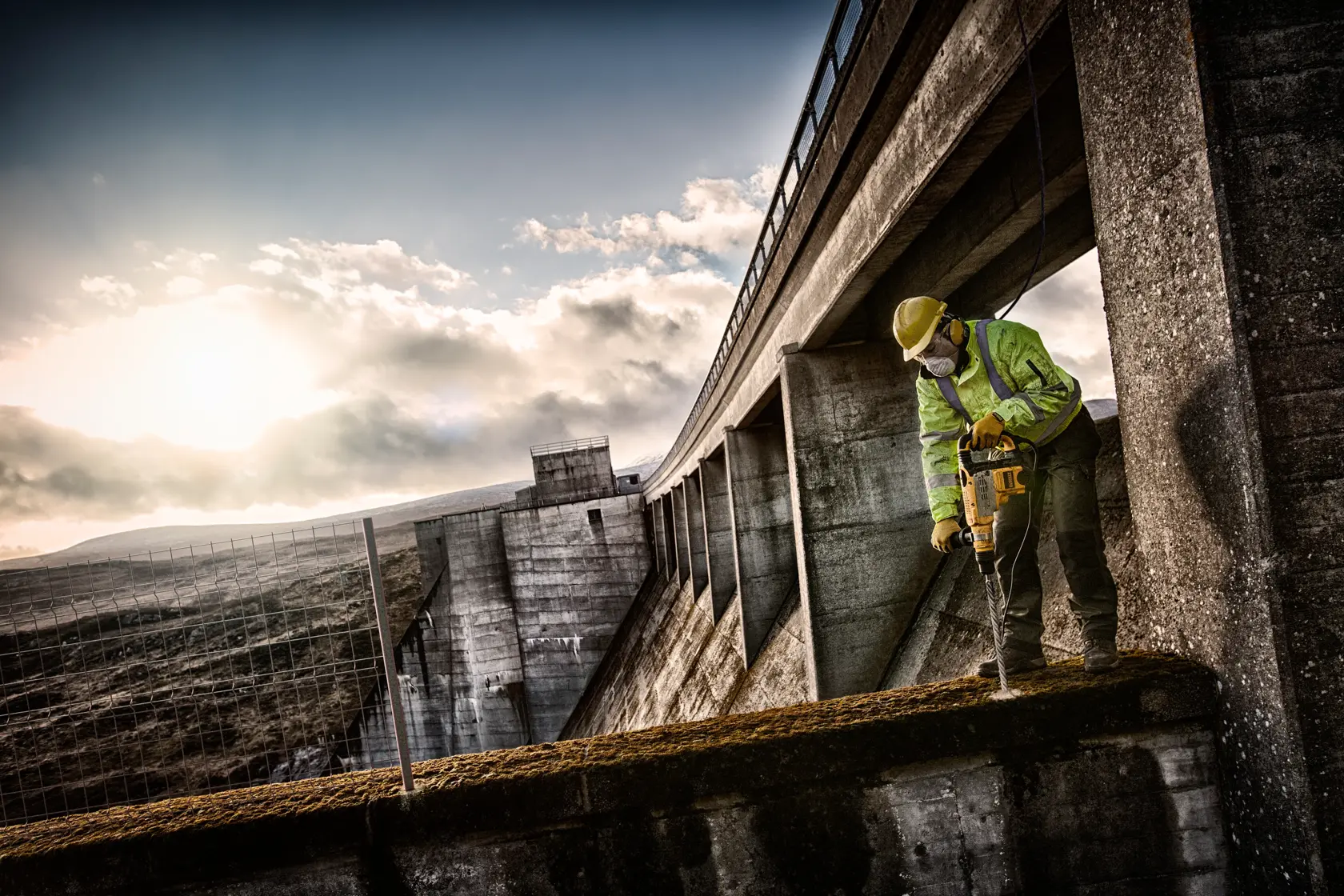 Arbeiter nutzt DEWALT-Bohrer auf Beton an Industriestandort, mit Sicherheitskleidung und Helm bei Sonnenlicht.
