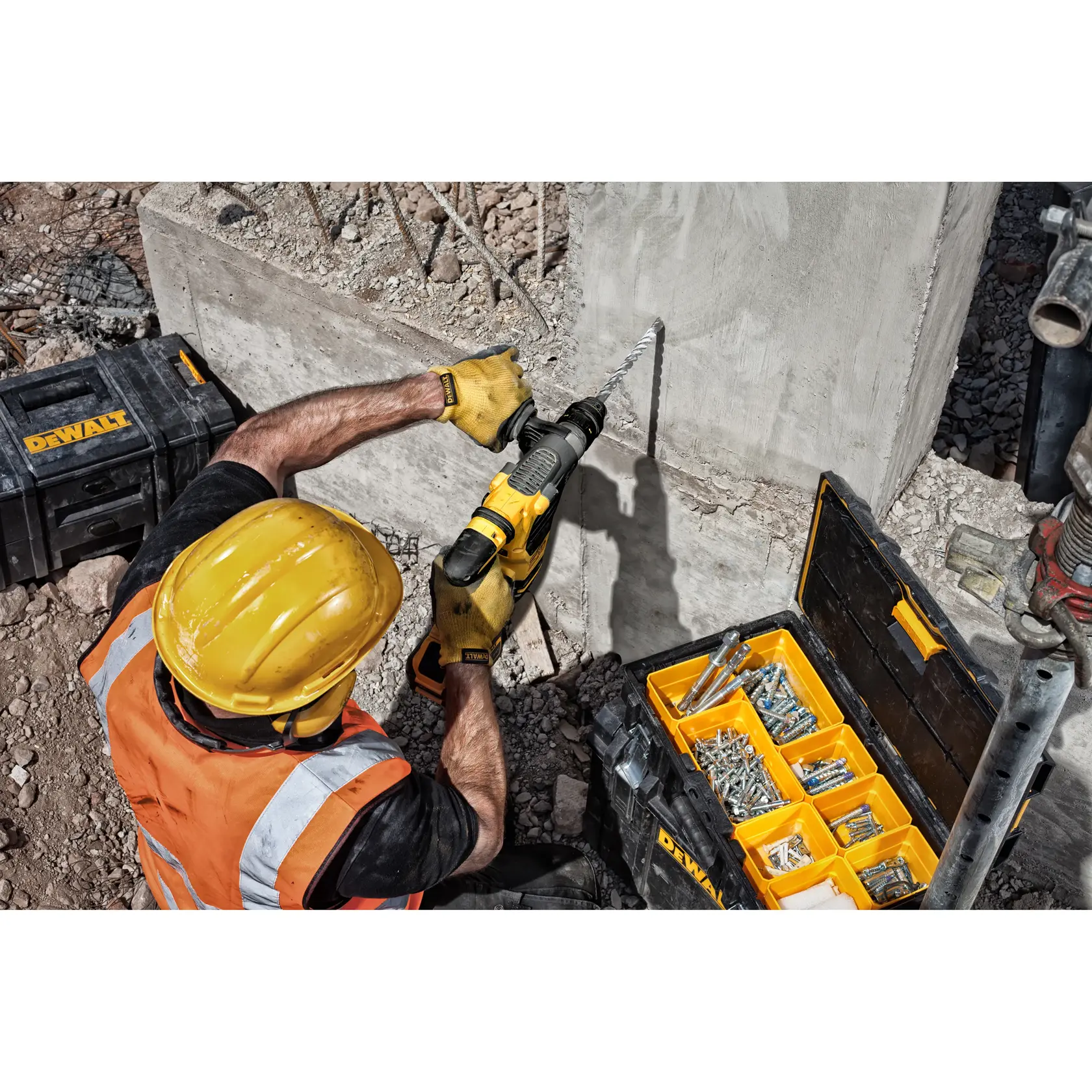 Trabajador con casco perfora muro de concreto con DEWALT; caja abierta con tornillos y brocas cerca.