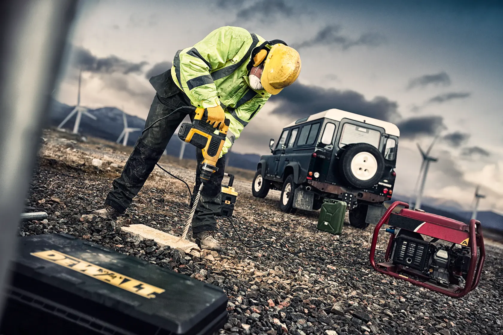 Worker drills into ground with DEWALT tool. Off-road vehicle and generator are nearby, wind turbines in background.