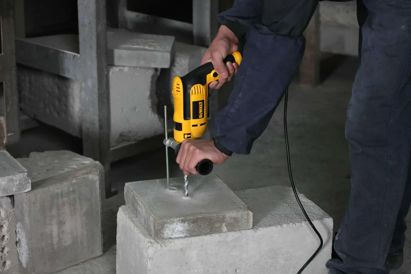 A person is using a yellow DEWALT electric drill to drill a hole into a concrete block in an industrial setting.